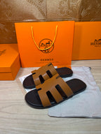 Hermes slide sandals, tan straps over black footbed, angled view on dust bag.