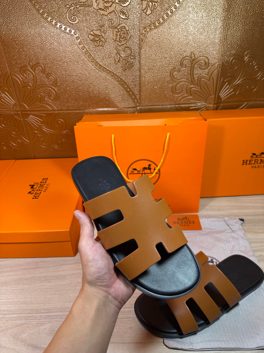 Hermes slide sandals held in hand, tan straps on black sole.