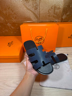 Hermes open-toe slide sandal, dark blue leather, held in hand with H-logo footbed.