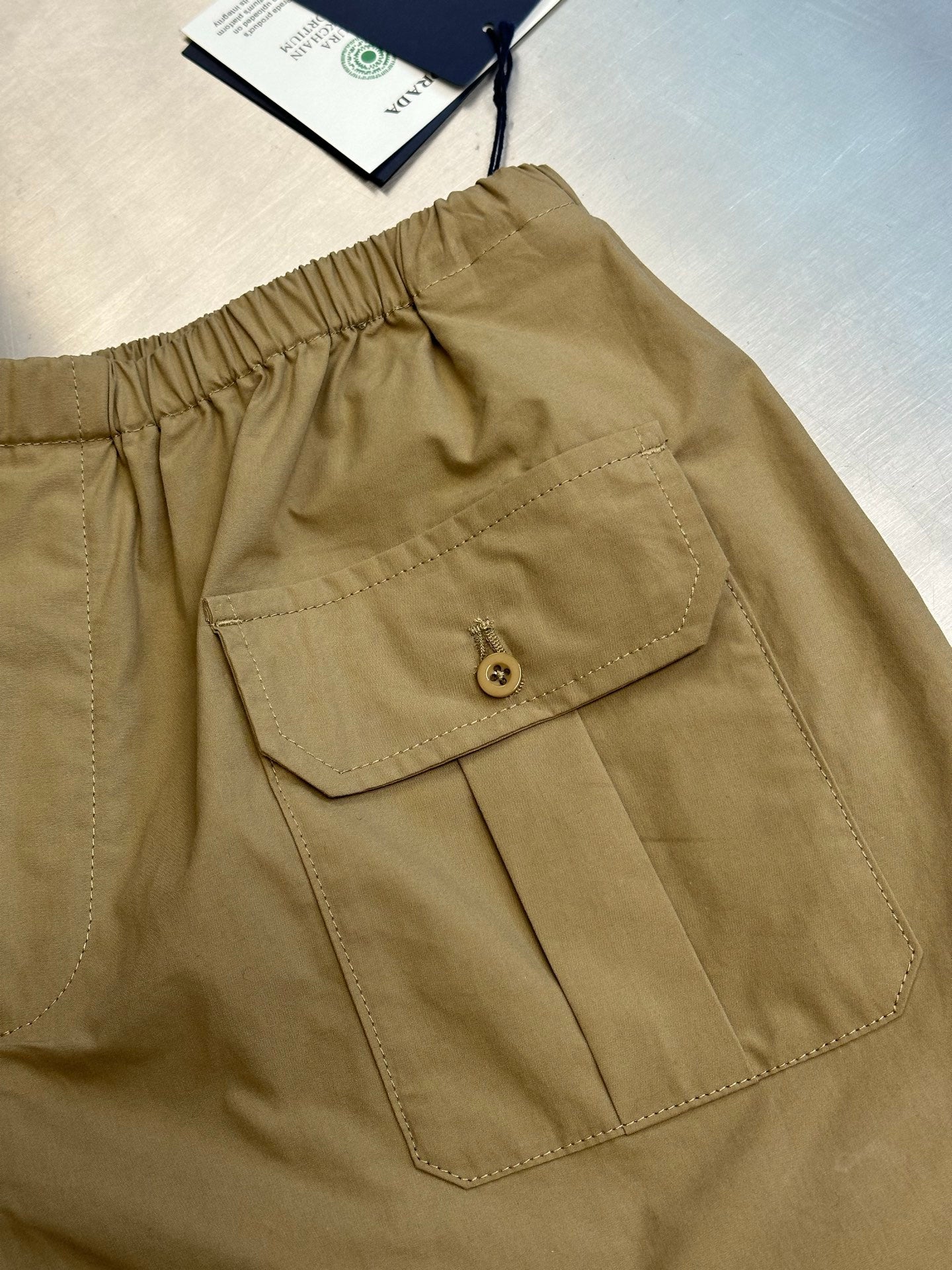 Beige cargo shorts with front patch pocket and round button closure.
