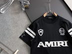 Black T-shirt front with AMIRI wordmark, crest emblem, and striped cuff details