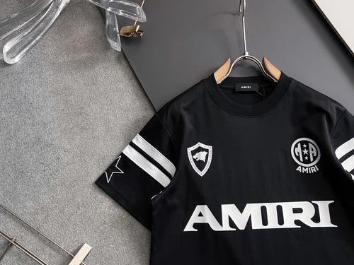 Black T-shirt front with AMIRI wordmark, crest emblem, and striped cuff details