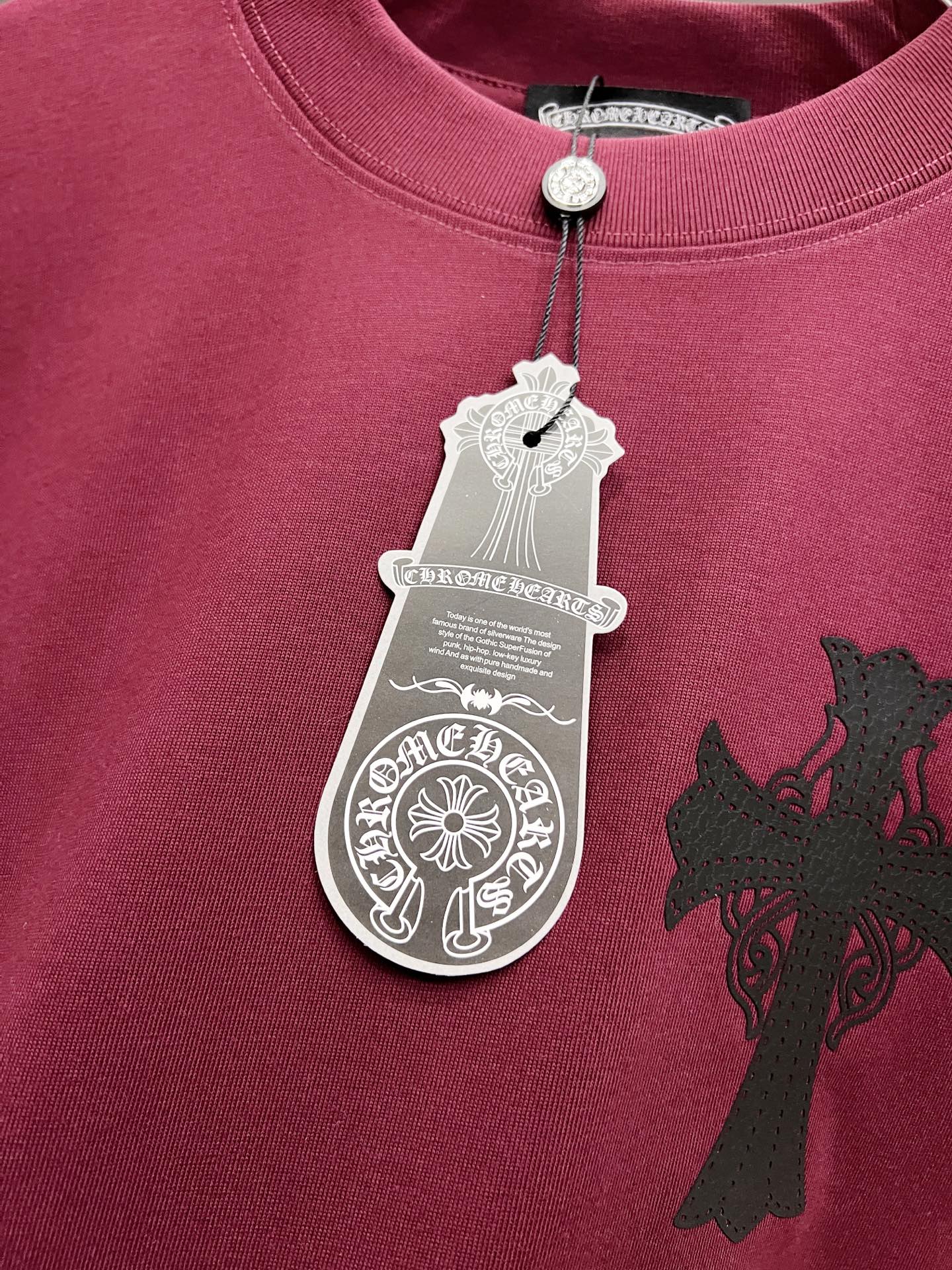Burgundy T-shirt with silver cross design and hanging white product tag at collar.