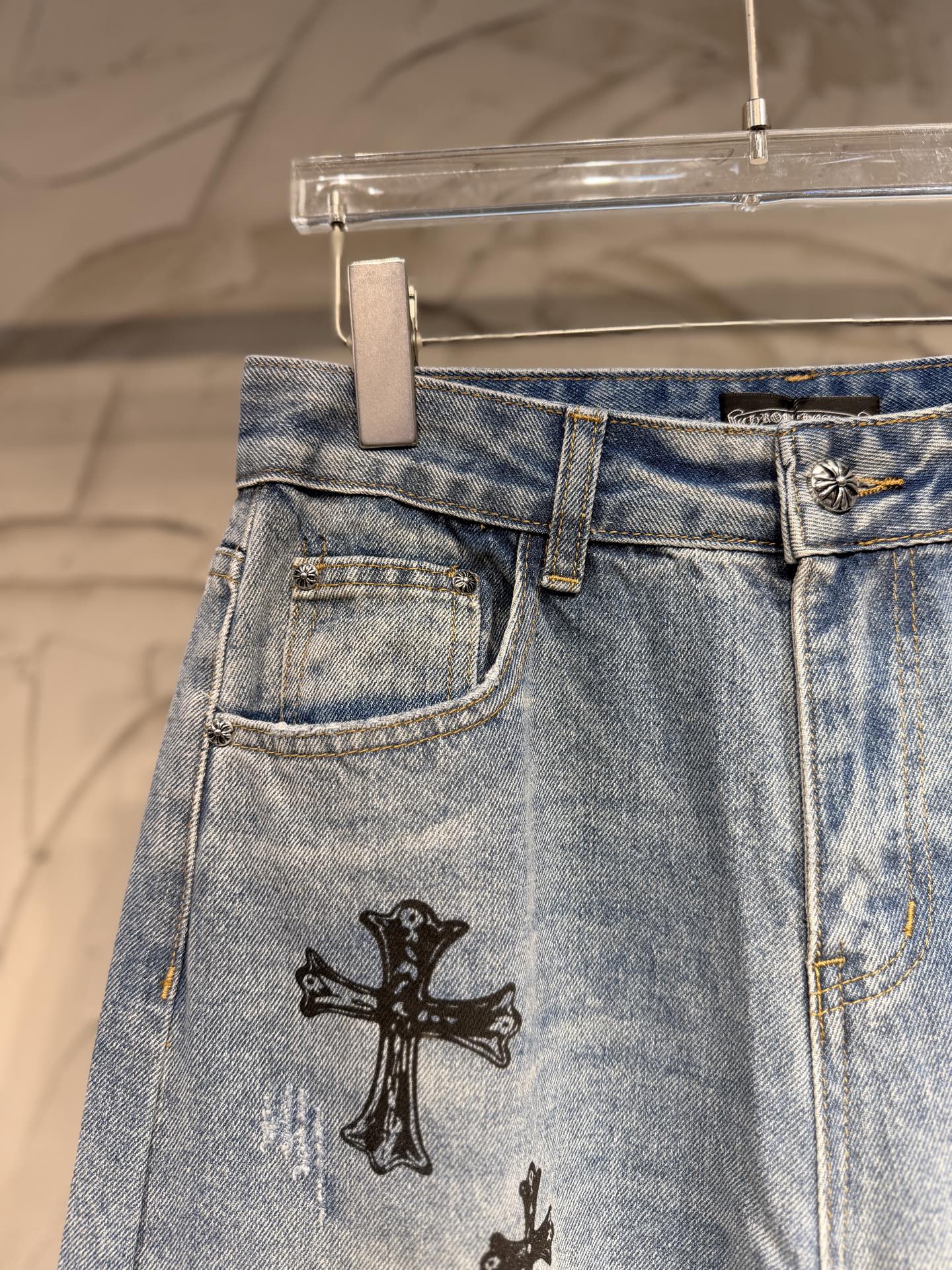 Light blue denim pants with button fly and black cross patch on the hip