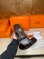 Brown leather slide sandals shown in hand with buckled front straps