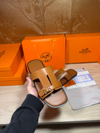 Hermes tan leather sandals held up, open toe and front buckle strap detail.