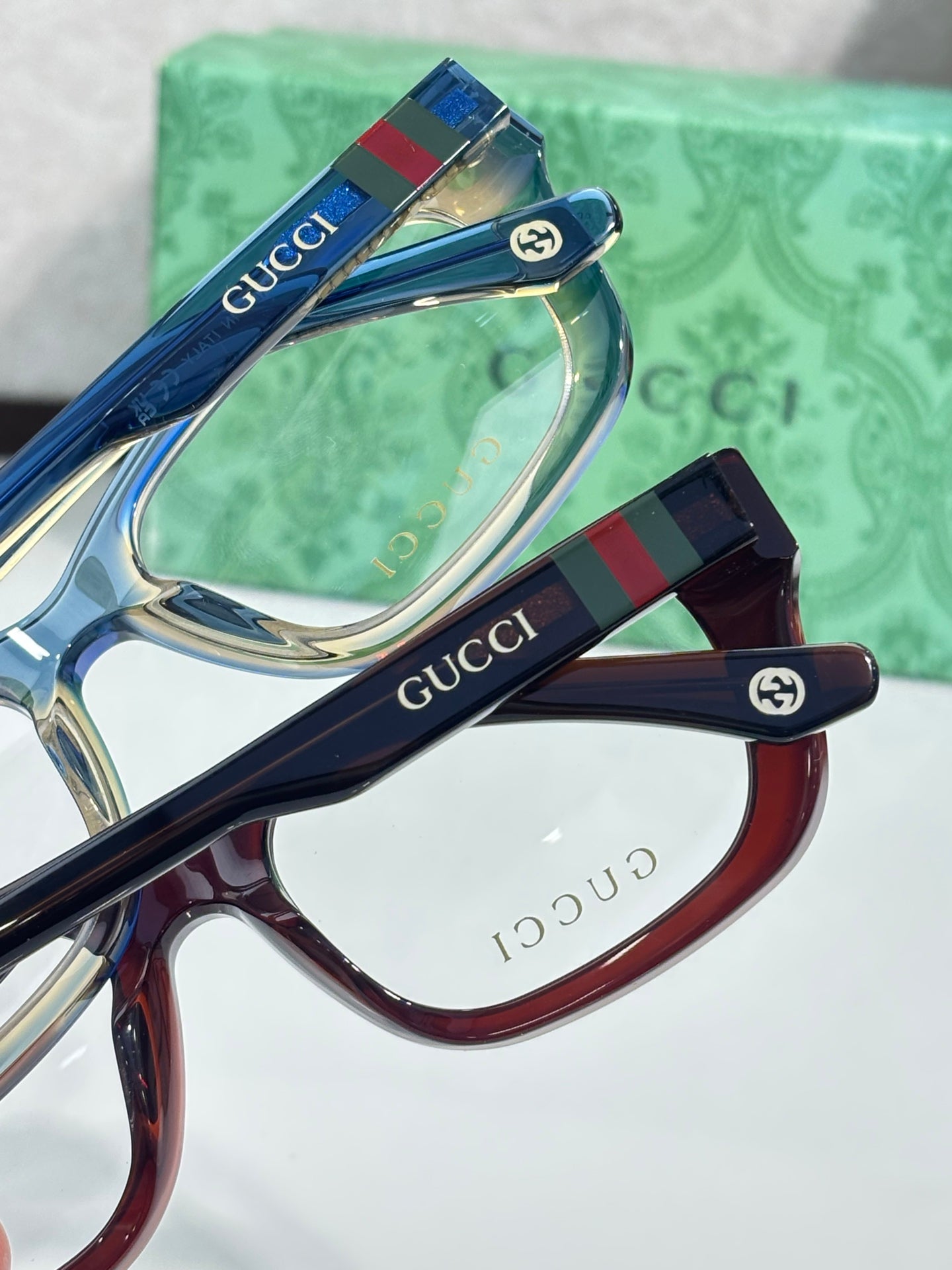 Gucci eyeglasses close-up: glossy temple logos with clear lenses on blue and maroon frames.