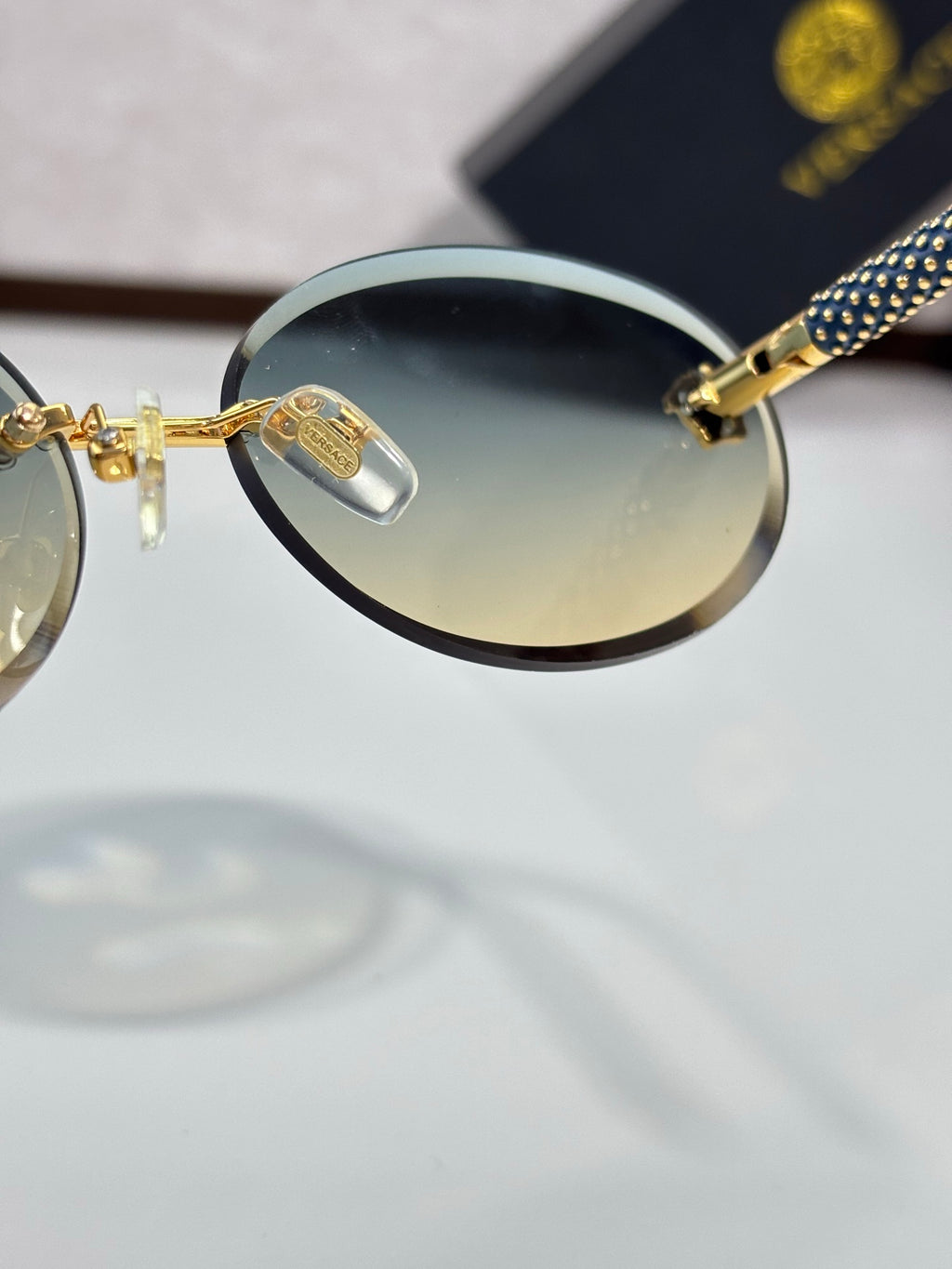 Versace sunglasses close-up: round tinted lens, gold bridge, blue-and-black temple.