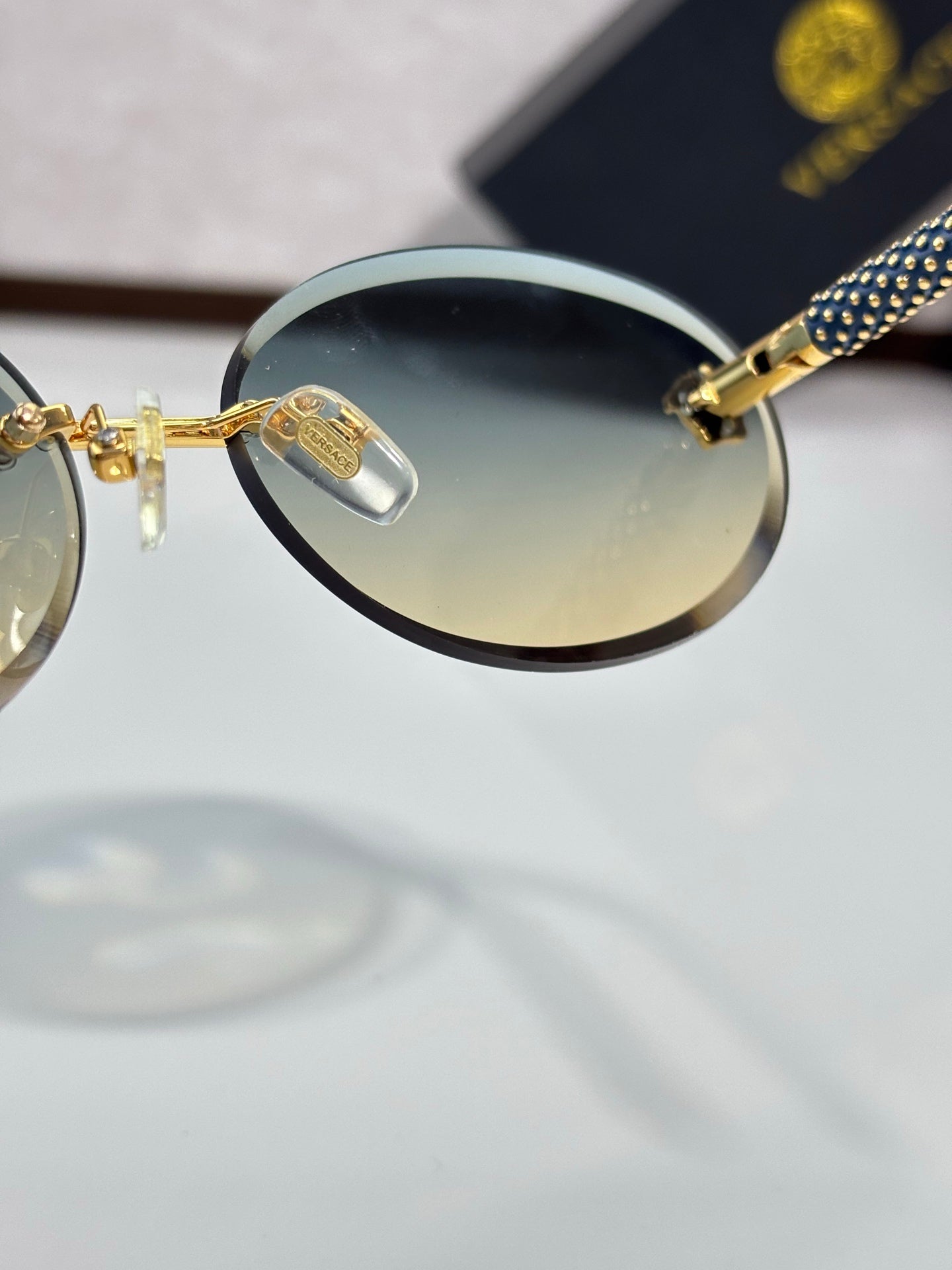 Versace sunglasses close-up: round tinted lens, gold bridge, blue-and-black temple.