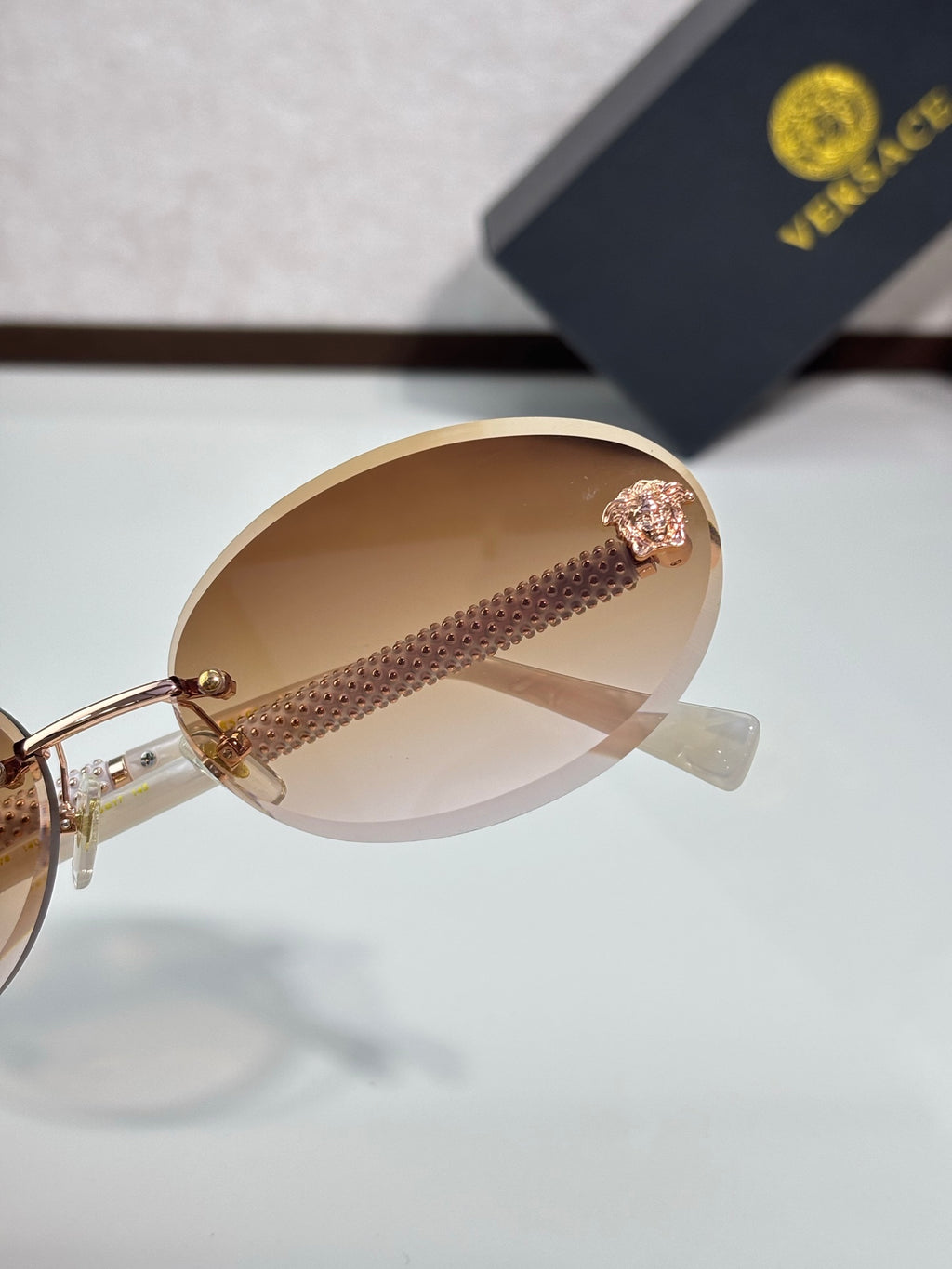 Versace sunglasses close-up: round tinted lens, gold temple detail, pink emblem.