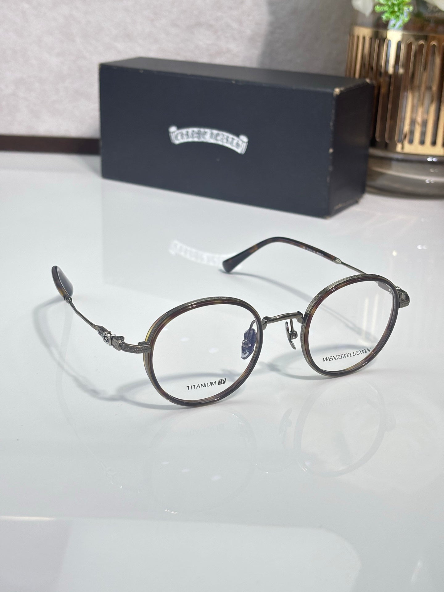 Round eyeglasses with dark frames, clear lenses, and gold-toned decorative temples.