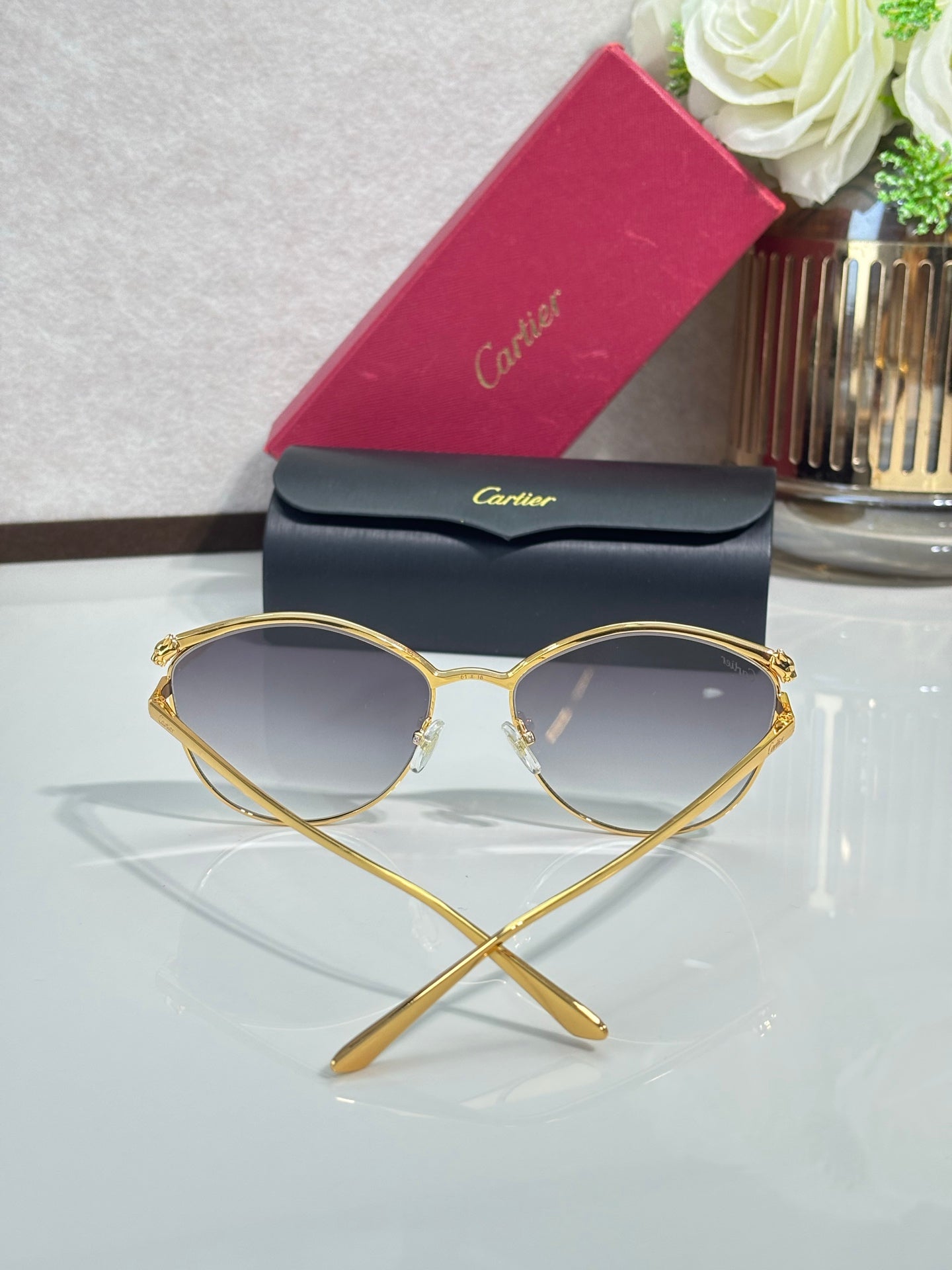 Gold round sunglasses with tinted gray lenses on clear stand, with red protective case.