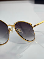 Close-up of gold round sunglasses frame with dark tinted lenses and gold temple detail.