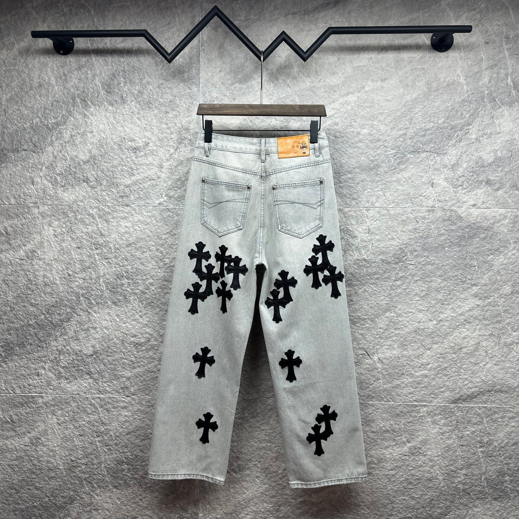 Light wash denim pants with black cross appliqués and tan leather patch at waistband