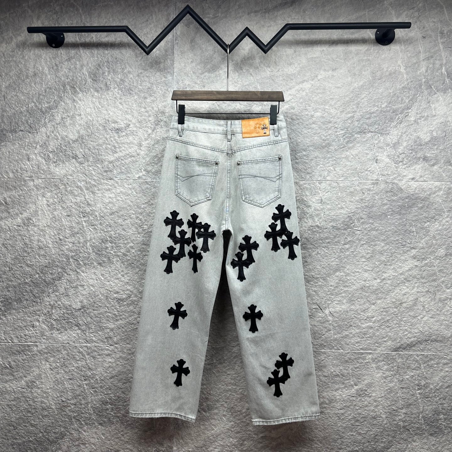 Light wash denim pants with black cross appliqués and tan leather patch at waistband
