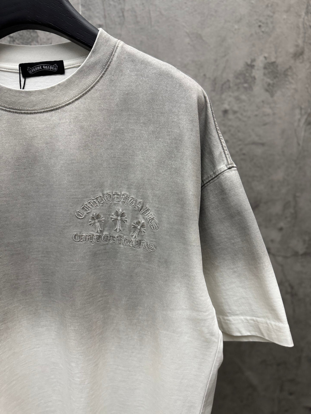 Light gray T-shirt front close-up with small circular chest embroidery and sleeve seam