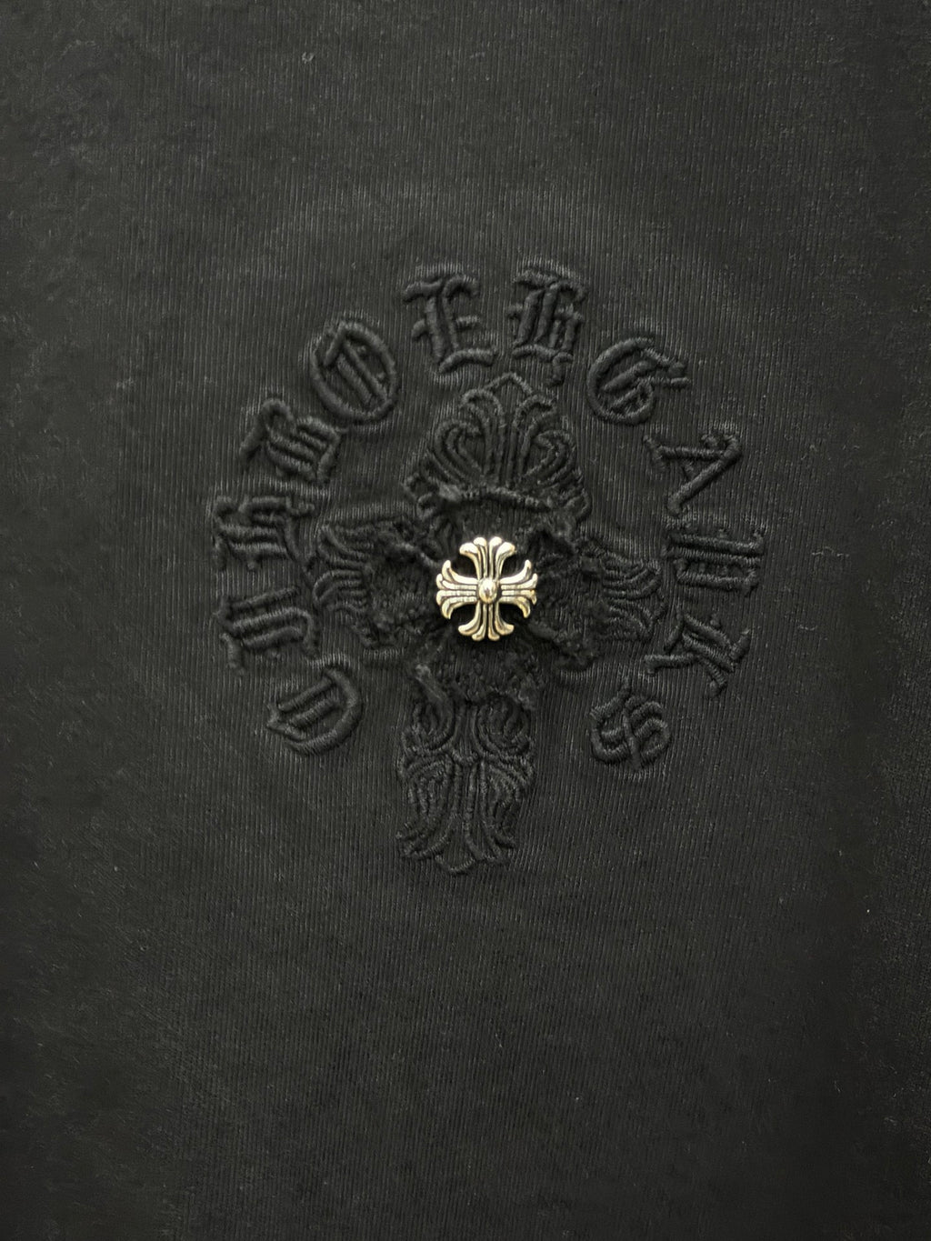 Black fabric T-shirt with detailed embroidered cross emblem and gold center detail.