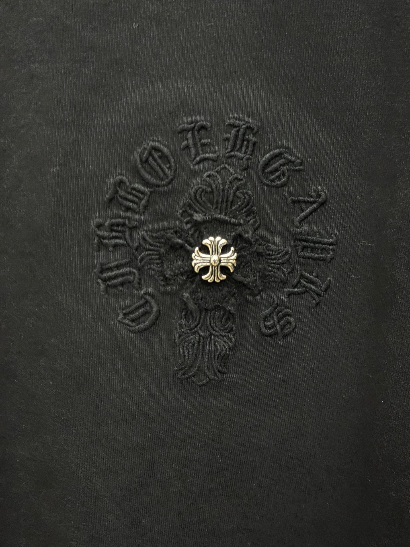 Black fabric T-shirt with detailed embroidered cross emblem and gold center detail.