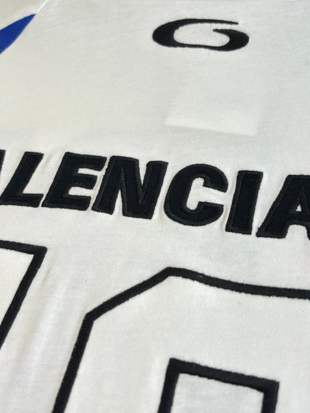 Balenciaga logo graphic on white jersey front, close-up of black lettering and blue stripe