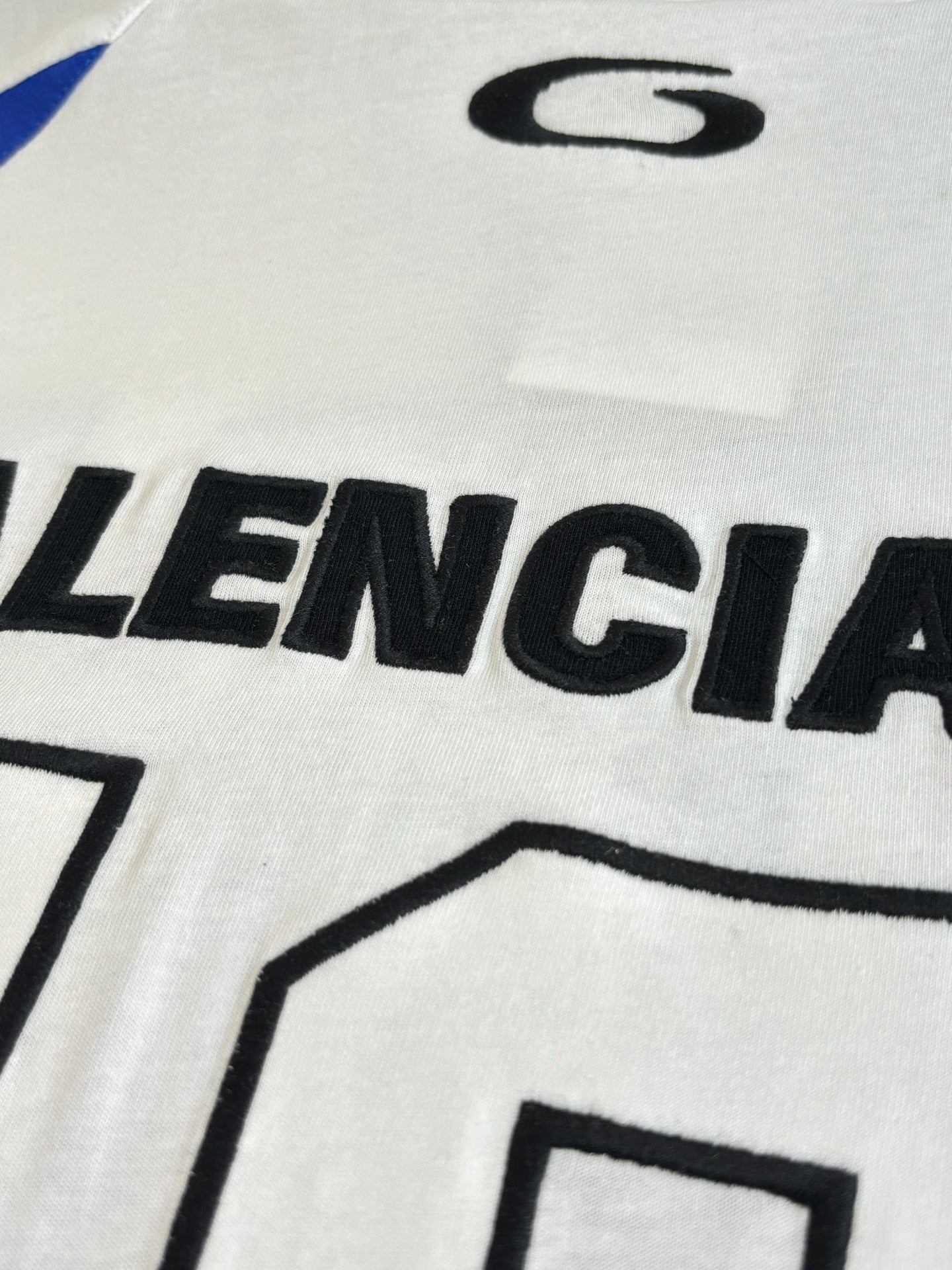 Balenciaga logo graphic on white jersey front, close-up of black lettering and blue stripe