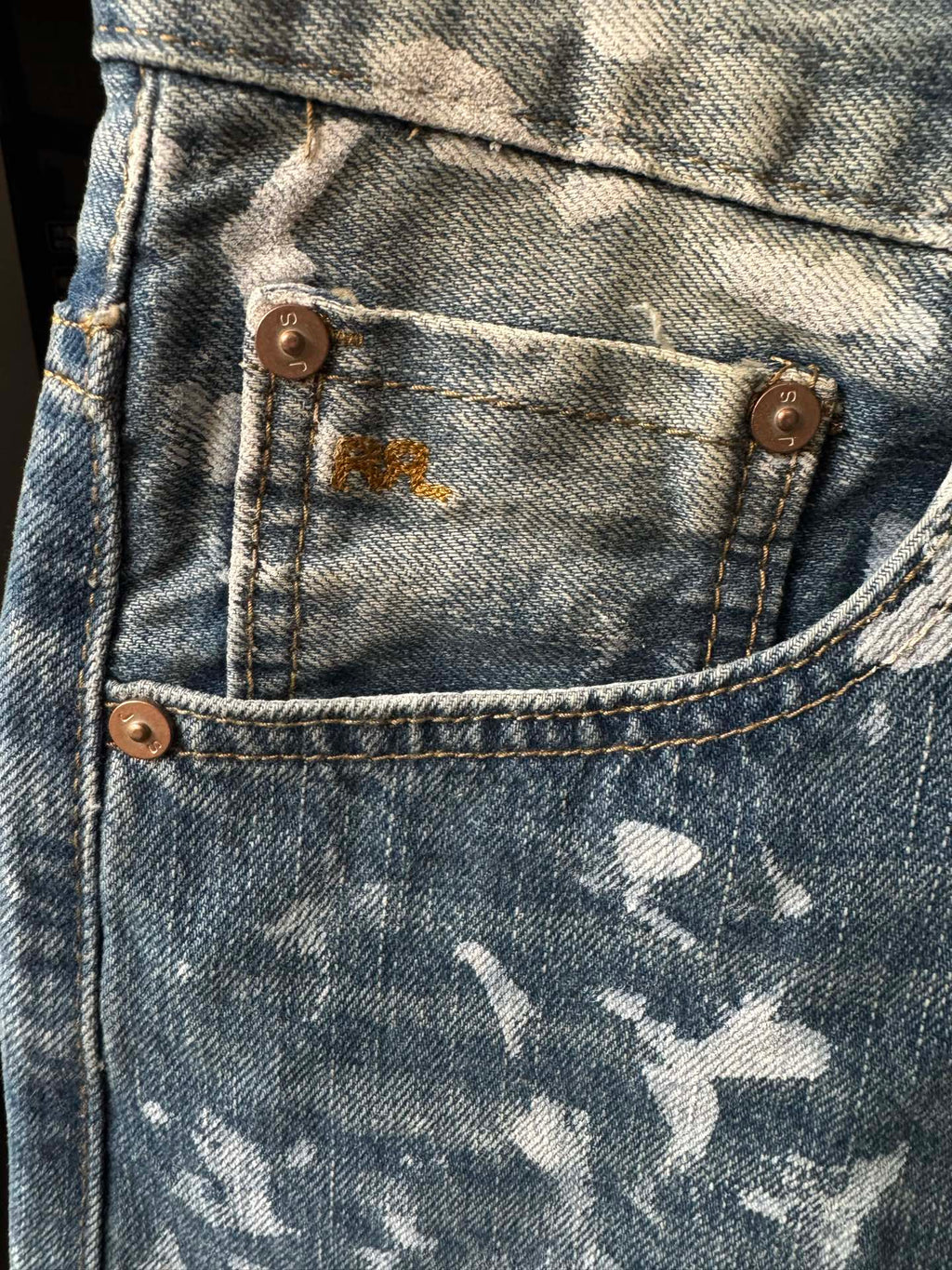 Light-wash denim jeans with bronze buttons and white speckle patches, close-up