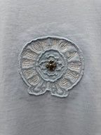 Light blue T-shirt fabric close-up of round embroidered emblem with small gold detail