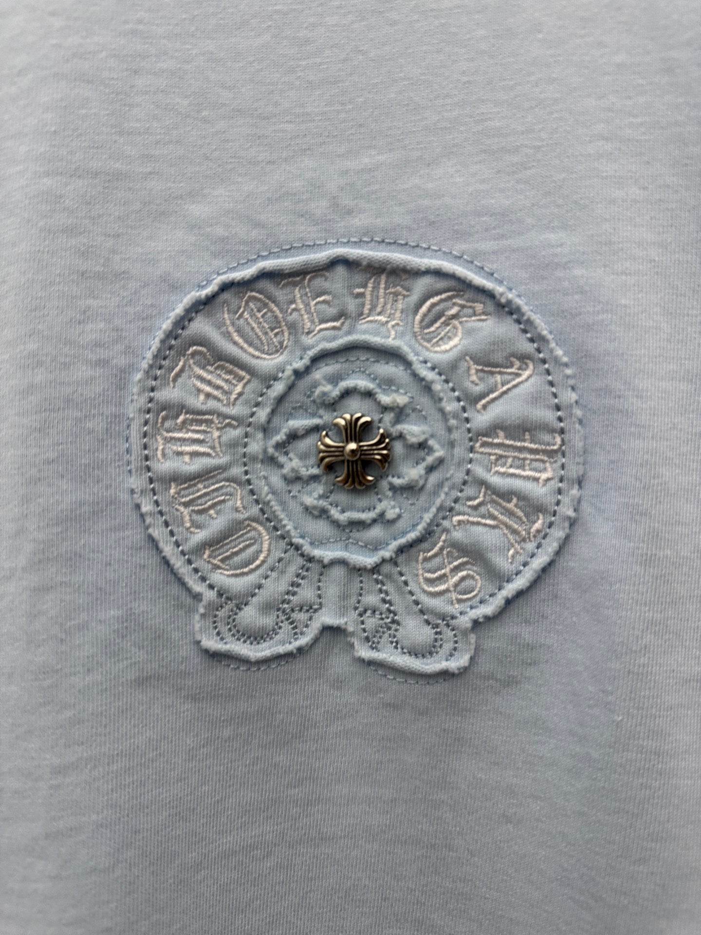 Light blue T-shirt fabric close-up of round embroidered emblem with small gold detail
