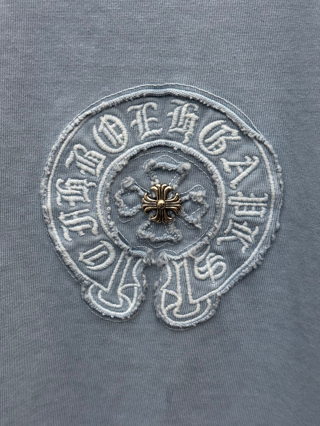 Round crest embroidery close-up on light blue T-shirt with gold center detail