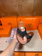 Hand holding a black leather thong sandal with H logo strap and tan sole.