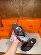 Hand holding brown leather flip-flop sandal, dark H strap on upper