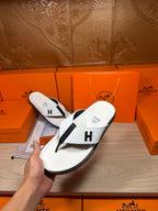 White thong sandal held by hand, black strap with H logo, with box behind