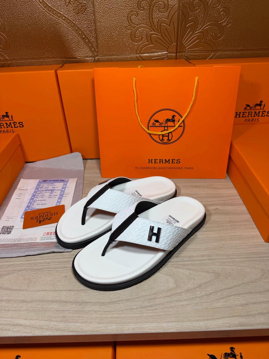 White thong sandals with black strap and H logo, angled view on display tray