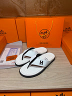 White thong sandals with black strap and H logo, angled view on display tray