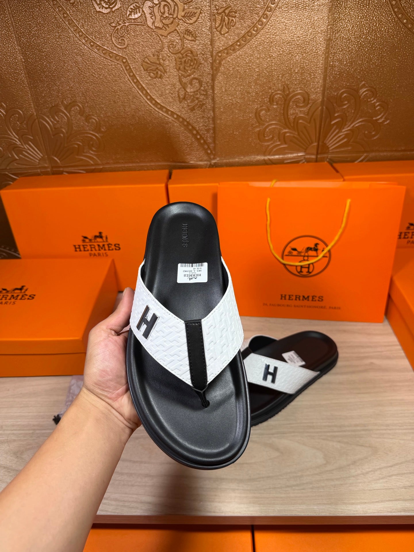 Hermes sandals held up showing black sole and white strap with H logo at toe.