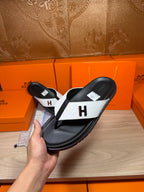 Hermes sandals held up: black sole and straps with white panel and H logo.