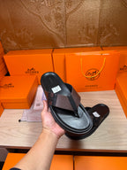 Hand holding black leather thong sandal with textured crossover strap and label