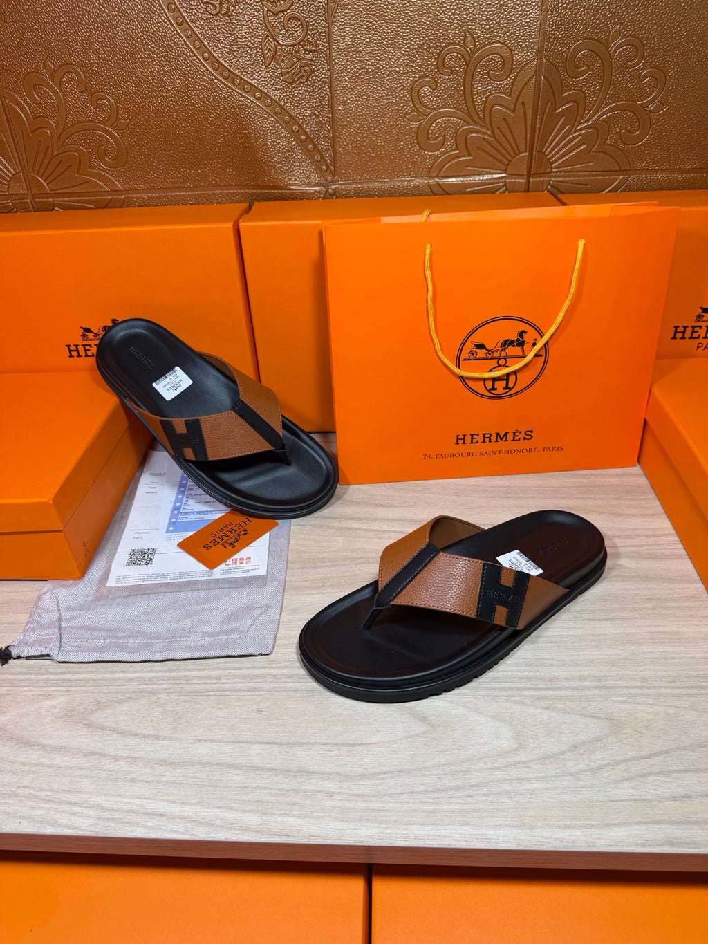 Black thong sandals with tan strap and H buckle detail, one laid flat