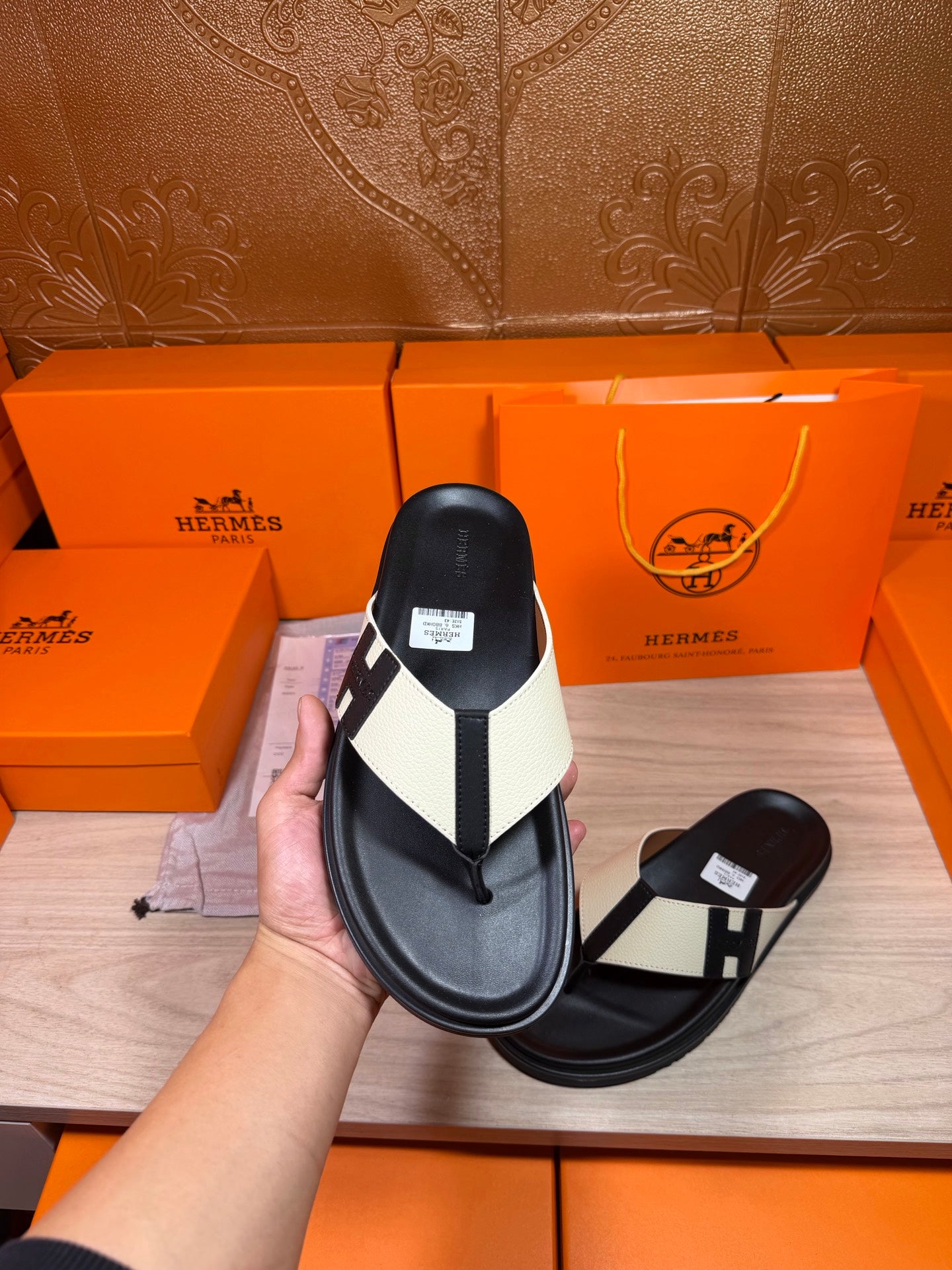 Hand holds black thong sandal showing white strap panels and square logo buckle