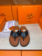 Black leather thong sandals with brown rubber sole, shown paired on display tray