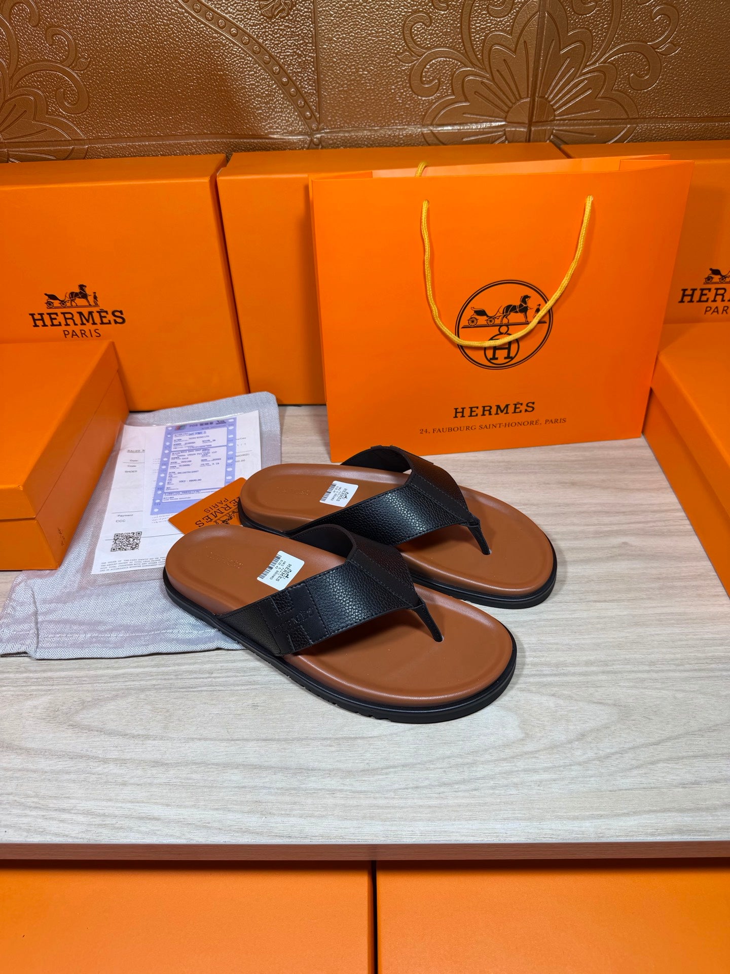 Black leather thong sandals with brown sole, angled view on display tray