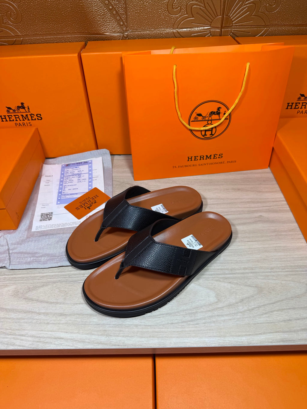 Black leather flip-flop sandals with brown sole, shown with box and tags nearby
