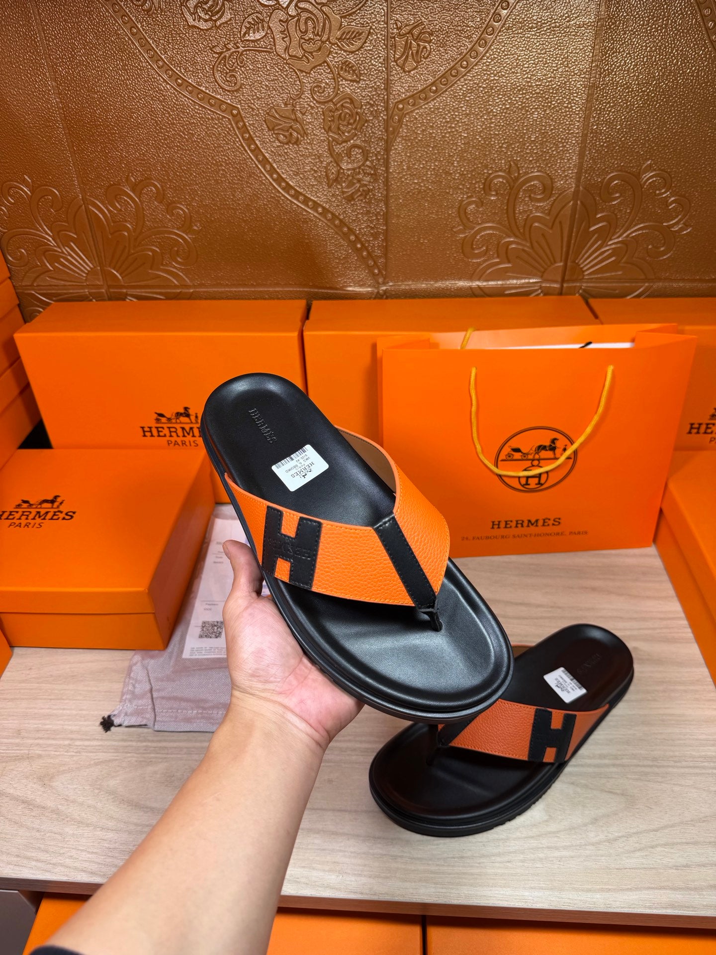 Hand holding black-and-orange H logo sandal with thong strap and footbed.