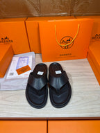 Hermes black leather slide sandals with cross strap and flat rubber sole.