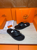 Hermes black leather slide sandals with cross strap and thick flat outsole.