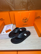 Hermes black leather slide sandals with textured cross strap and flat sole.