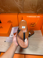 Brown leather thong sandal held in hand with dark H strap and flat sole