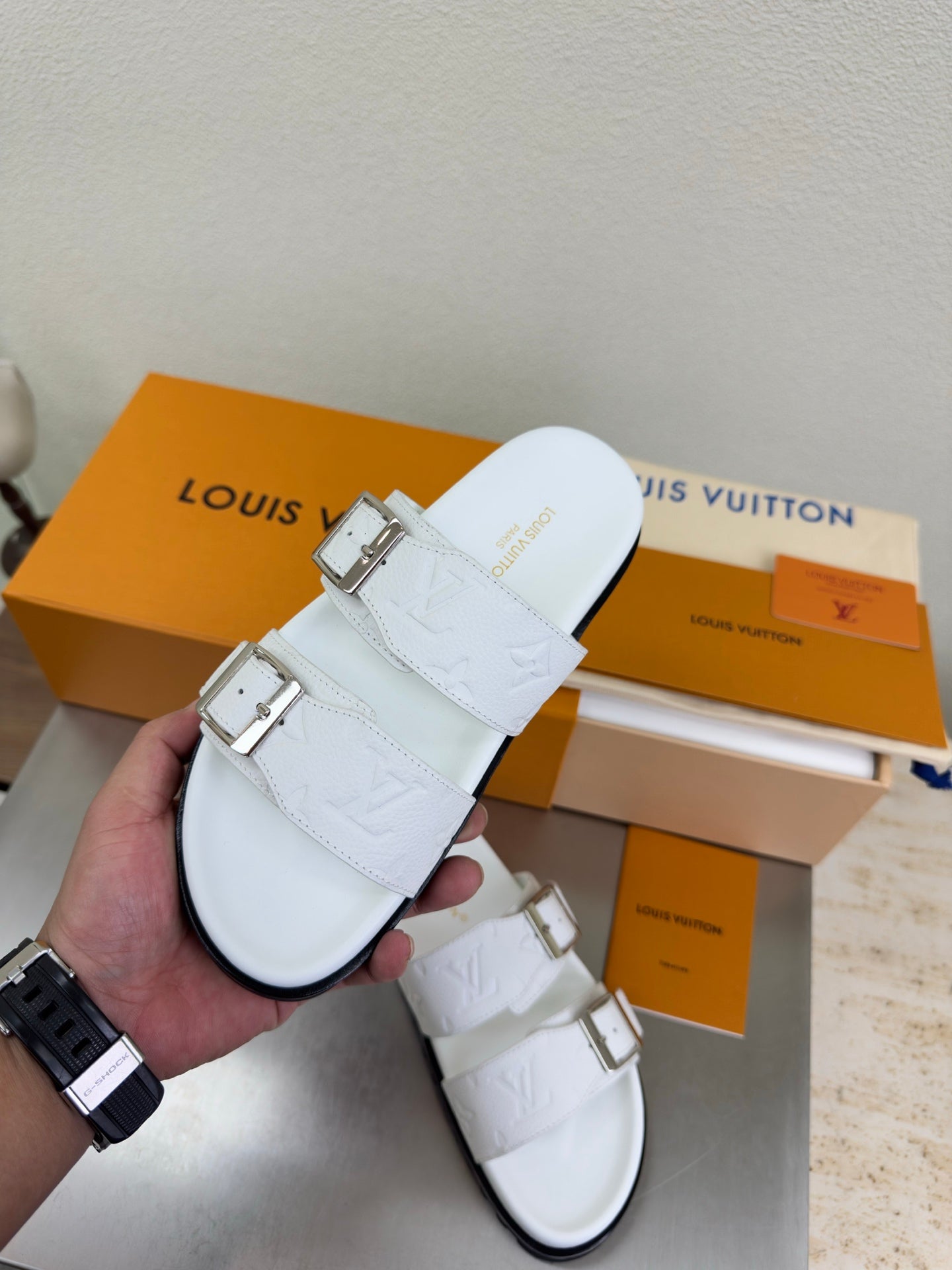White Louis Vuitton buckle sandals held in hand, embossed strap detail and silver tabs