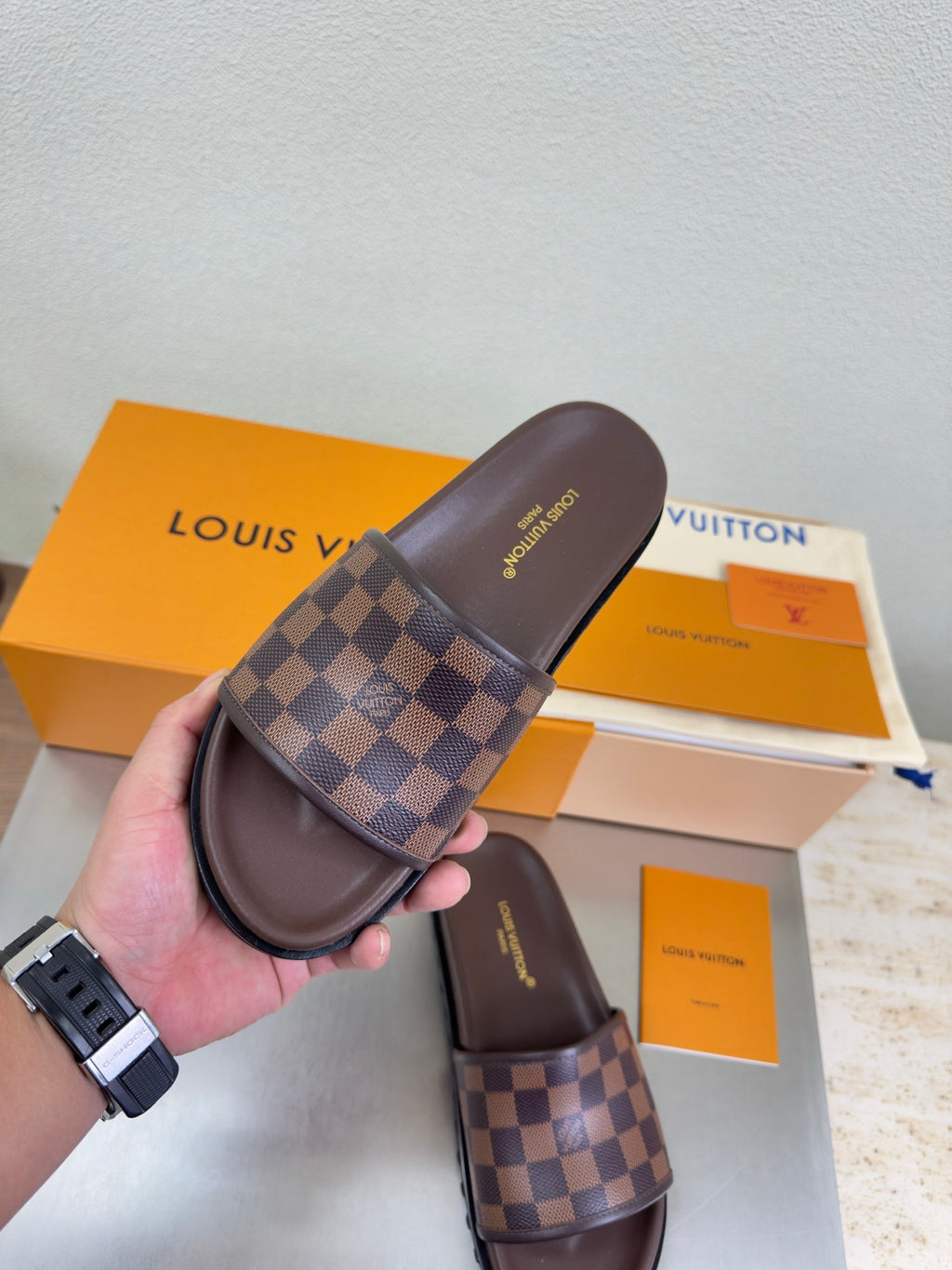 Louis Vuitton damier check slide sandal held in hand, brown upper with single strap.