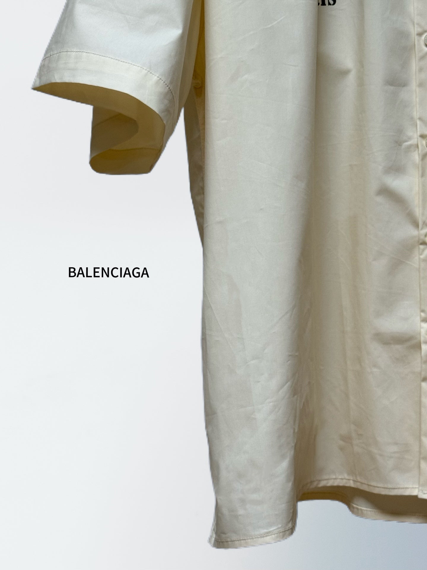 Balenciaga cream short-sleeve shirt, curved hem with button front and partially visible sleeve cuff.