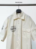 Balenciaga cream short-sleeve button shirt with pointed collar, chest pockets, and front text.